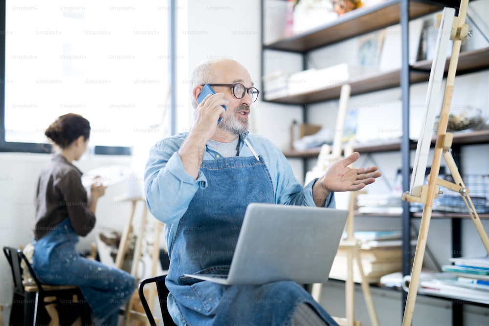 Mature bearded man in apron sitting in front of easel in workshop and talking to someone on smartphone