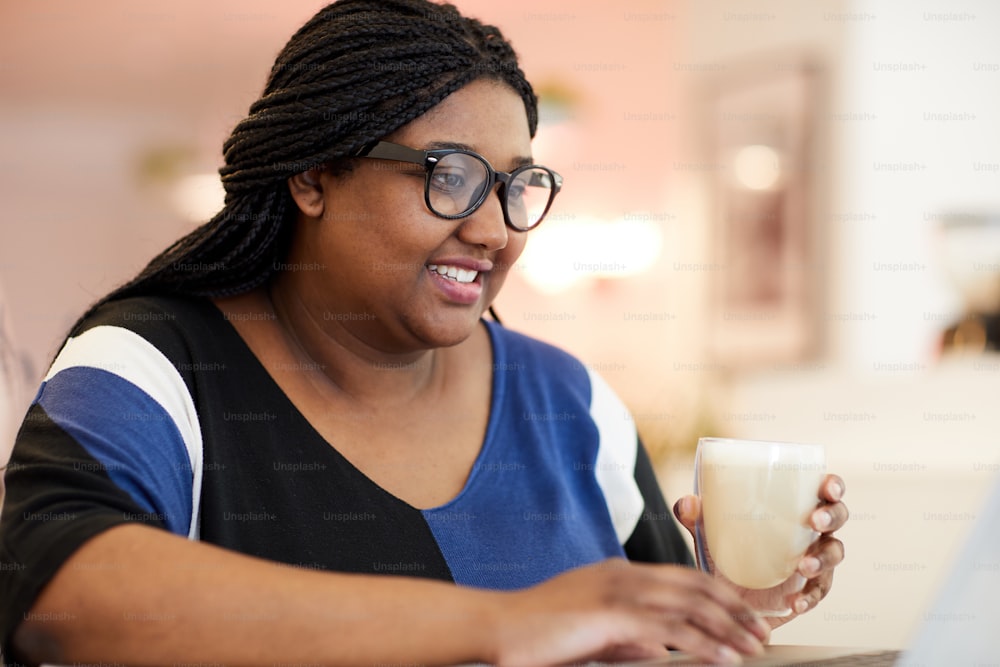 Young mixed-race woman having milkshake in cafe while sitting in front of laptop and searching for data