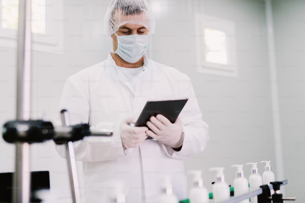 Bild eines jungen Mannes in steriler Kleidung mit Tablet . Steuerung der Produktionslinie in der Fabrik. Flaschen mit Kosmetikprodukten auf Produktionslinie.
