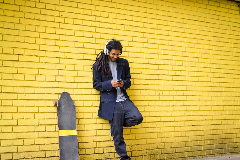 Young cute hipster dreadlocks man leaned on yellow wall listening music with headphones on his head with his long board near him.