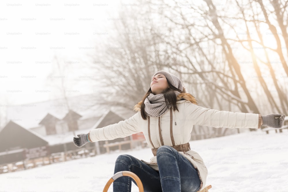 Beautiful young woman sitting on a sleigh with her eyes closed and arms spread, facing the sky and warm winter sun