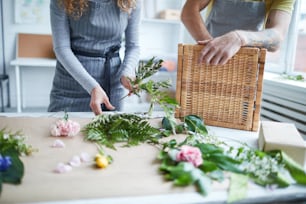 Young female florist putting fern leaves into bunch with her colleague standing near by