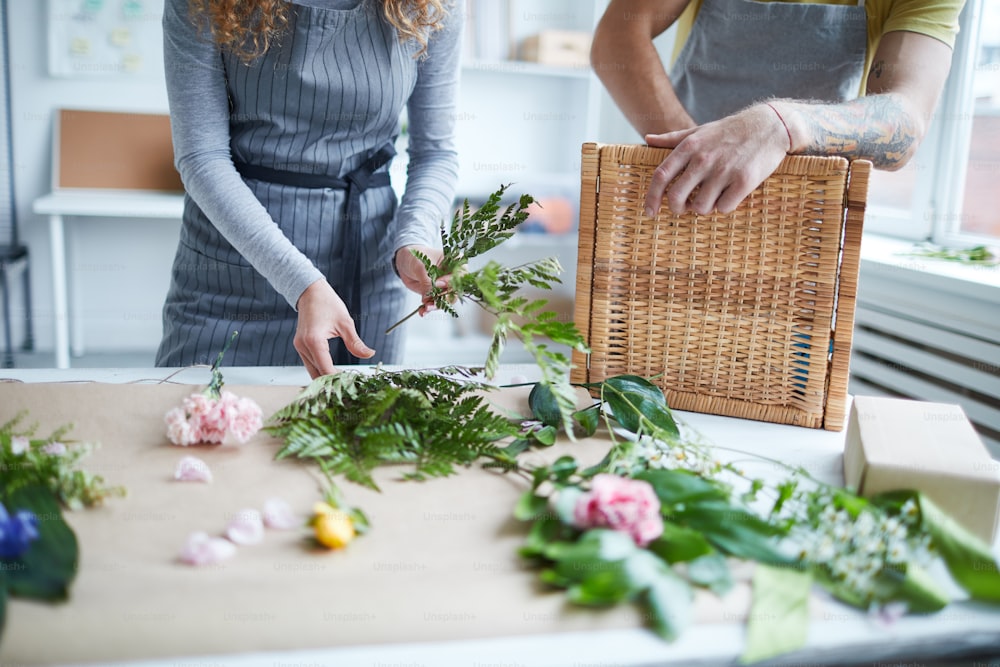 Young female florist putting fern leaves into bunch with her colleague standing near by