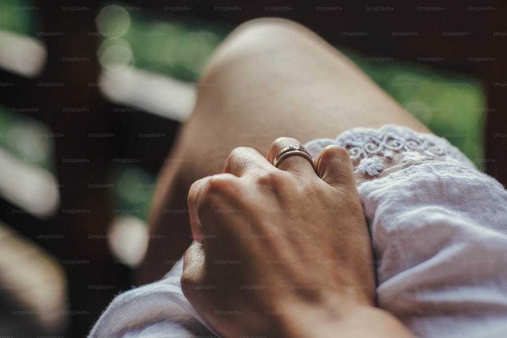 mano con un anillo de compromiso inusual y hermoso. Muchacha de la mano con anillo de plata con piedra en la pierna y vestido bohemio blanco, sentada en el porche de madera. Espacio para el texto