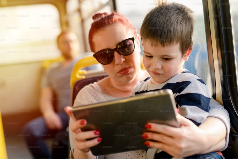 バスの中で息子と一緒に座っている若い思いやりのある母親。男の子は彼女の膝の上に座ってタブレットを見ています。