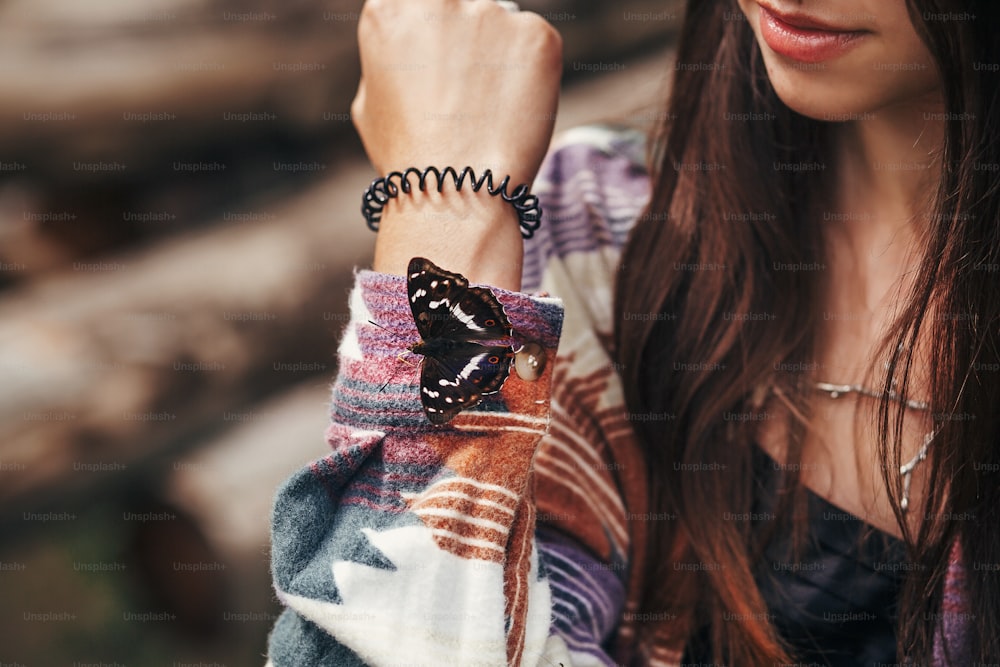 女の子の手に美しい蝶。スタイリッシュなヒップスター、山の森で指にアパチュラアイリスを握る帽子をかぶった幸せな女性。旅行と放浪癖のコンセプト。テキスト用のスペース。アメージング・モーメント