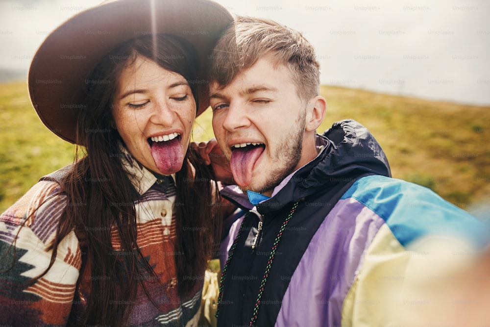 happy hipster couple making selfie and showing colored tongues from blueberries on top of sunny mountains. family having fun and smiling. summer vacation. space for text.