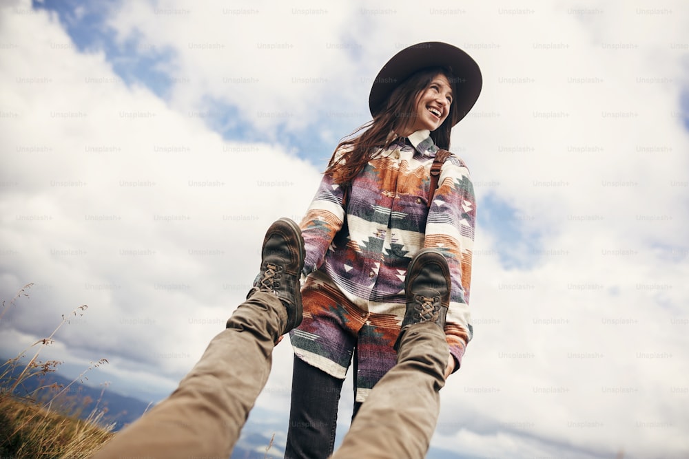 happy stylish traveler girl in hat holding man legs, pulling them and laughing, funny moment on top of sunny mountains. travel and wanderlust concept. space for text. couple traveling