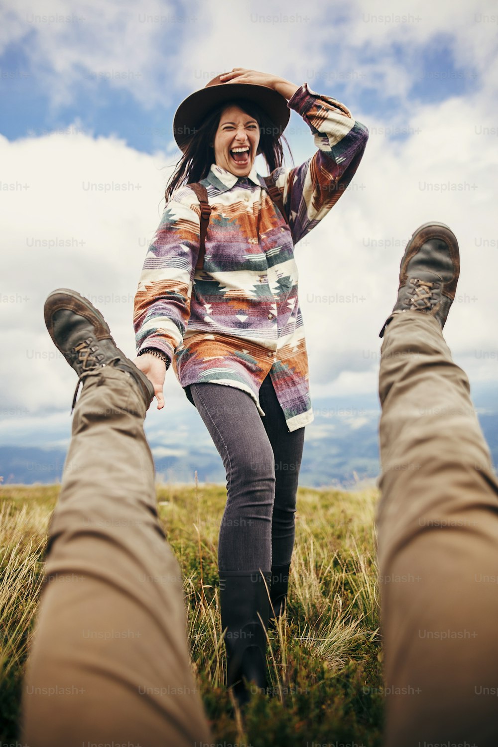 happy stylish traveler girl in hat holding man legs and laughing, funny moment on top of sunny mountains. travel and wanderlust concept. space for text. couple traveling