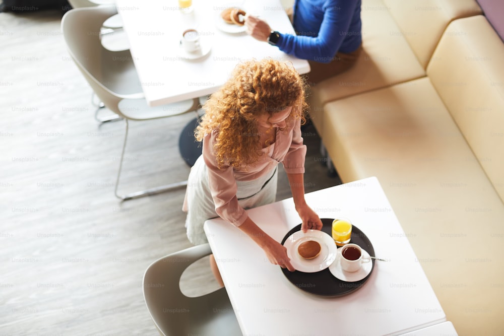 Direkt über Blick auf die lockige junge Kellnerin, die am Tisch steht und den Teller mit Pfannkuchen auf das Tablett stellt, arbeitet sie im Café