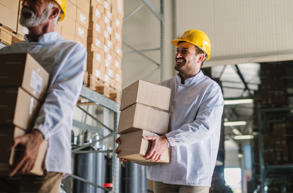 Picture of two male warehouse workers with helmets on their heads carrying boxes in their hands. Smiling and walking.
