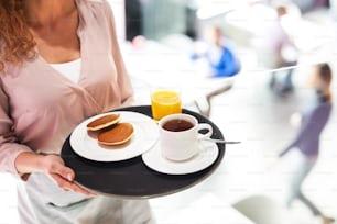 Close-up of unrecognizable waitress carrying tray with breakfast consisting of pancakes, coffee and orange juice, work in cafe