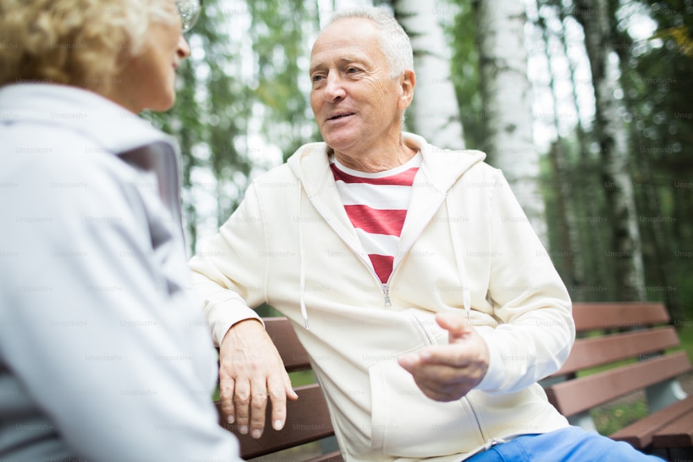 Mature man in activewear and his wife sitting on bench in park and having talk