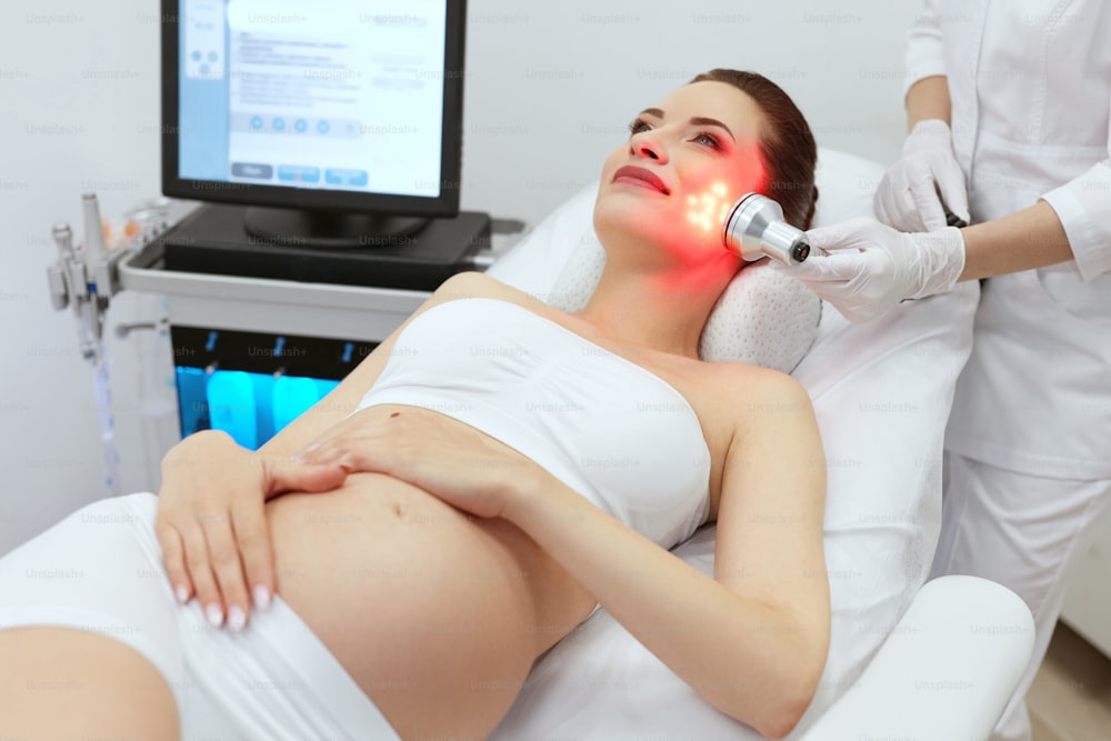 Cosmetology. Pregnant Woman Doing Red Light Therapy At Beauty Clinic. Cosmetologist Doing Facial Treatment. High Resolution