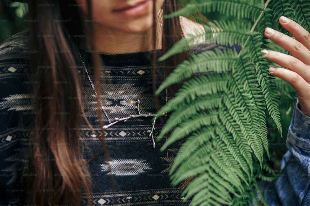 stylish hipster girl sitting in fern bushes, among fern leaves in forest. close-up of branch silver necklace. environmental concept. space for text. atmospheric moment. nature protection
