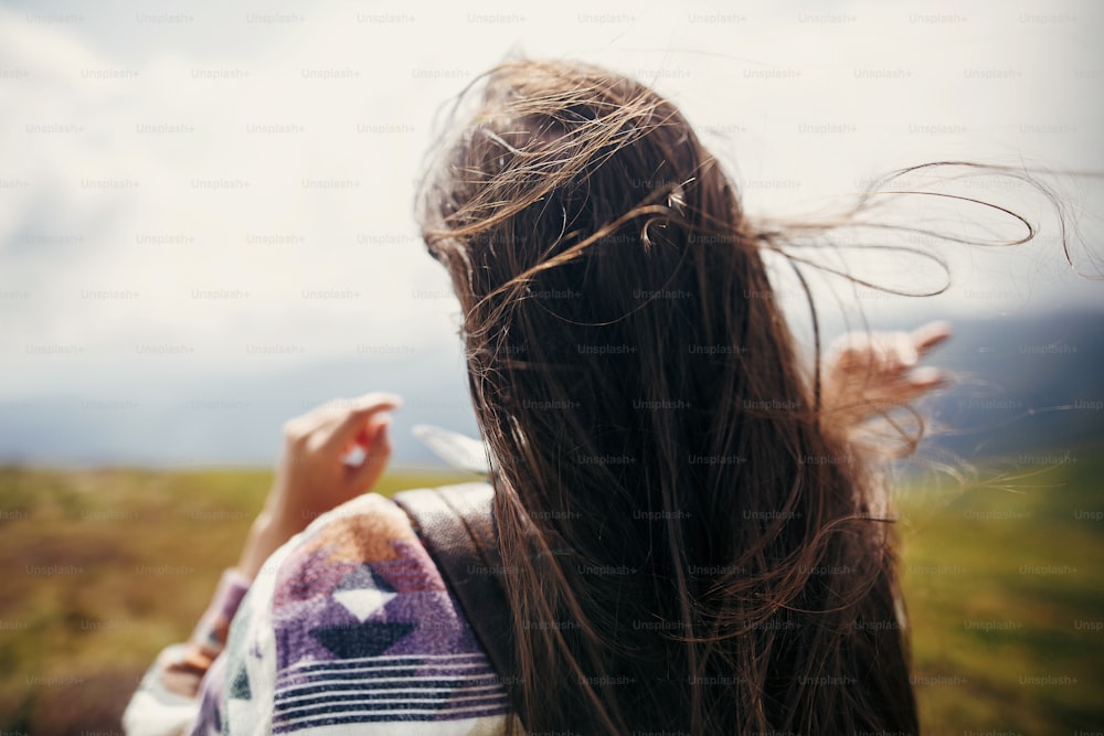 Reisendes Hipster-Mädchen mit windigen Haaren und Rucksack, auf sonnigen Bergen stehend. Platz für Text. Stilvolle Frau winkt mit Haaren. atmosphärischer Moment. Reise- und Fernwehkonzept.