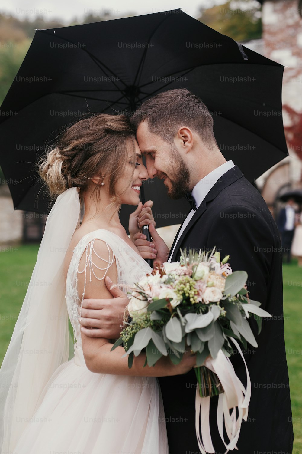 Gorgeous bride and stylish groom gently hugging under umbrella in rainy outdoors. Sensual wedding couple embracing. Romantic moments of newlyweds. Modern wedding photo