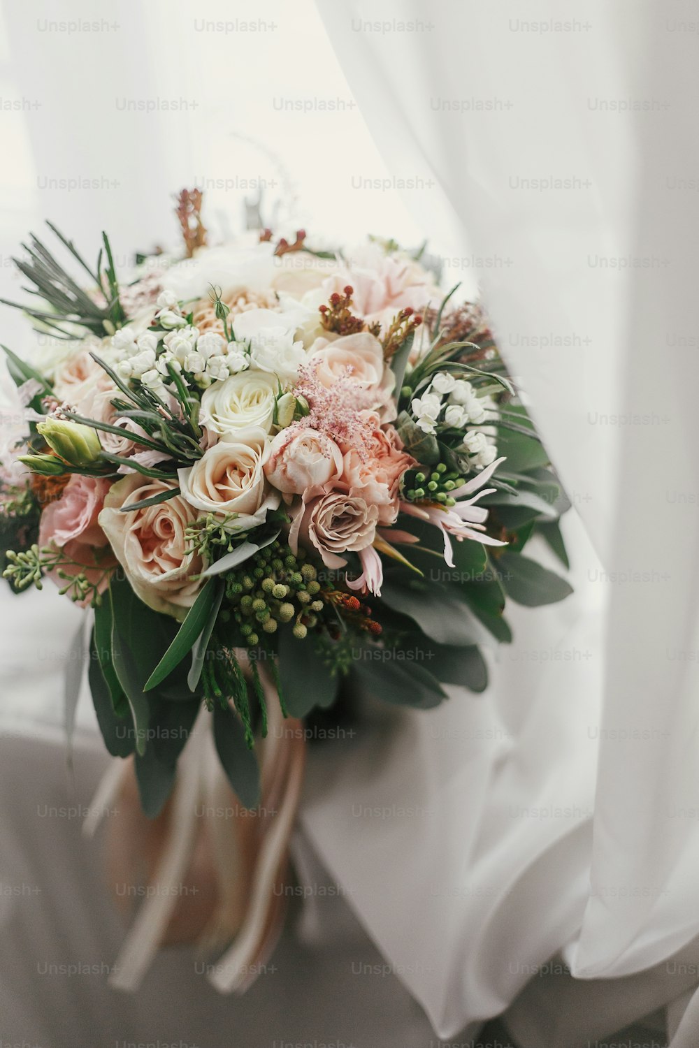 Stylish wedding bouquet of pink roses and green eucalyptus on background of window. Modern bride's bouquet on soft fabric in morning light. Wedding arrangements and decor