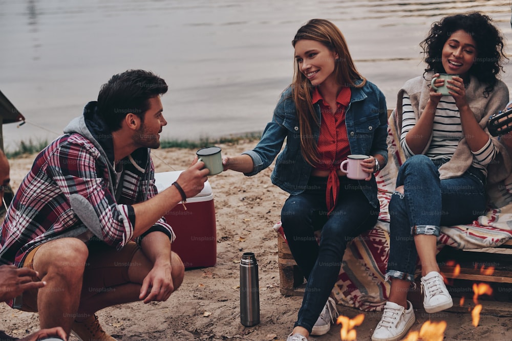 Group of young people in casual wear smiling and holding mugs while camping near the bonfire