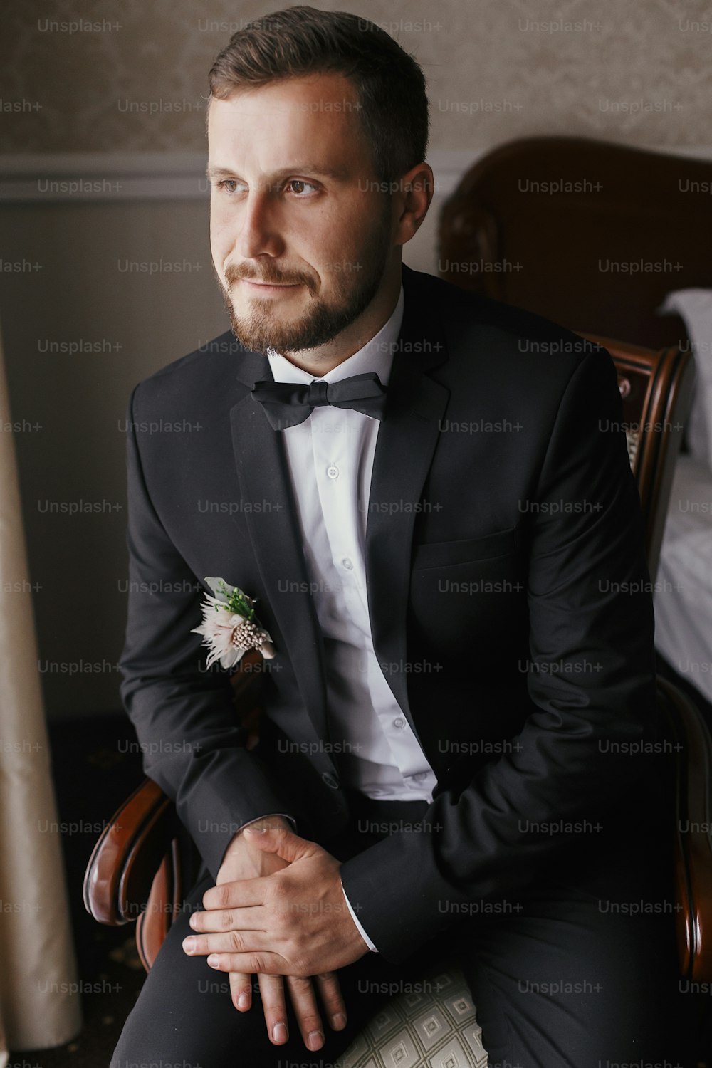Stilvoller Bräutigam in schwarzem Anzug und Fliege, sitzt im Stuhl bei Fensterlicht. Selbstbewusstes und fröhliches Porträt des Menschen. Bräutigam bereitet sich morgens vor