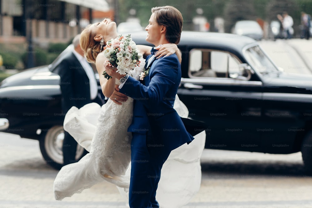 luxury wedding couple dancing at old car in light. stylish bride and groom hugging and embracing in city street. romantic sensual moment. woman looking at man