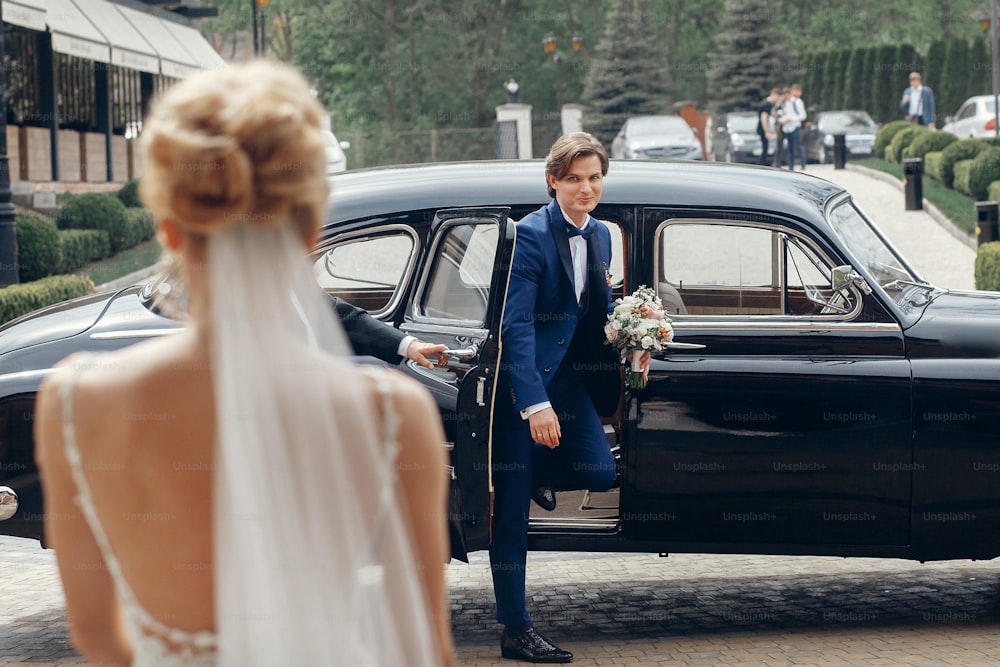 車から歩いていく幸せな花婿を見ているスタイリッシュな花嫁。豪華な結婚式のカップルは、街の通りで抱擁、ファーストルック。ロマンチックで情熱的な官能的な瞬間。