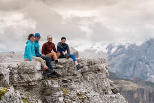 ドロミテの山頂の棚に座り、ランコフェルとマルモラーダの山頂の素晴らしい景色を見ている4人の若い男女のハイカー