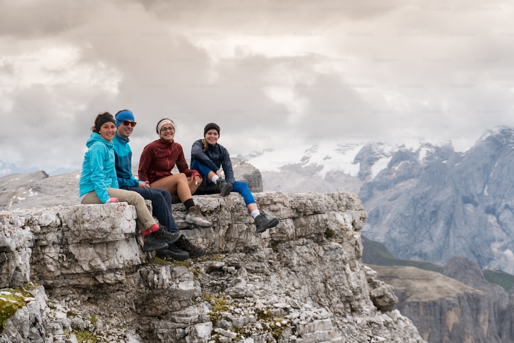 vier junge Wanderer und Wandererinnen sitzen auf einem Bergvorsprung in den Dolomiten und betrachten die herrliche Aussicht mit Langkofel und Marmolada