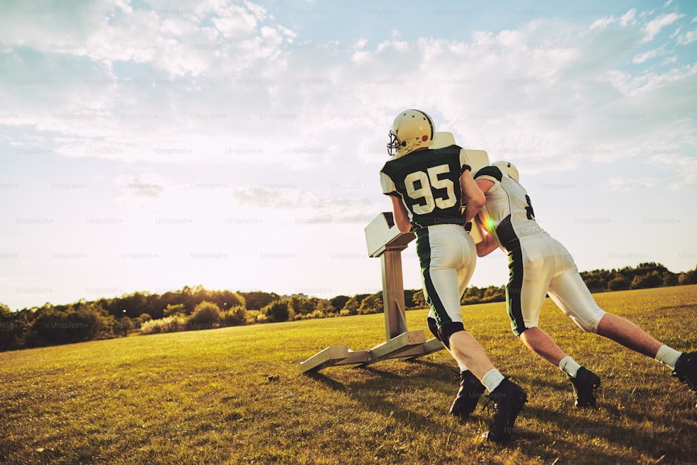 Dois jogadores de futebol americano praticando tackles com um trenó de tackle do lado de fora em um campo esportivo no final da tarde