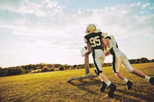 Two American football players practicing tackles with a tackle sled outside on a sports field in the late afternoon