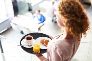 Young woman carrying pancakes, tea and juice on tray while moving to one of tables in cafe