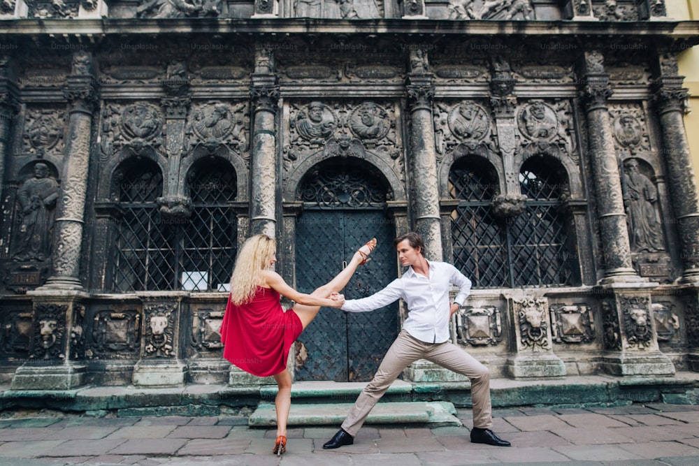 햇볕이 잘 드는 거리의 오래된 건물에서 포옹하고 춤을 추는 사랑에 빠진 아름다운 행복한 커플. 세련된 힙스터 신랑과 빨간 드레스를 입은 금발 신부는 춤을 추고, 전문 댄서들. 낭만적 인 순간들