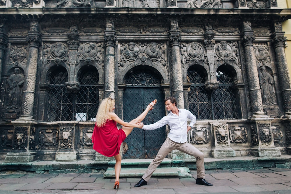 Schönes, glückliches verliebtes Paar, das sich im alten Gebäude in der sonnigen Straße umarmt und tanzt. Stilvoller Hipster-Bräutigam und blonde Braut im roten Kleid tanzen, professionelle Tänzer. Romantische Momente
