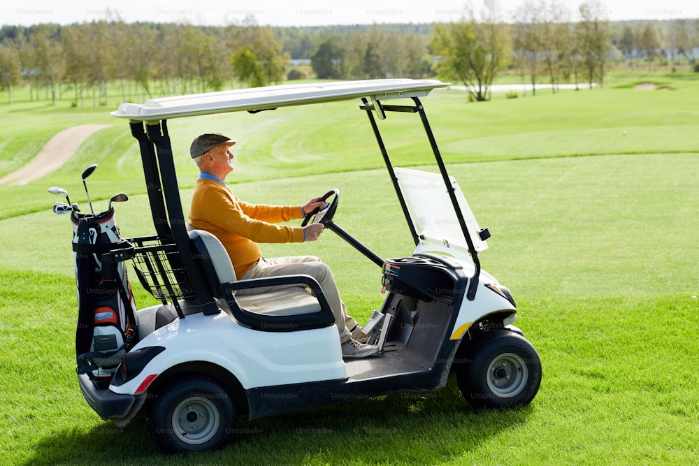 Homem envelhecido contemporâneo dirigindo carro de golfe ao longo do vasto campo verde enquanto vai para o jogo