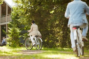 Rear view of young couple in casual clothing riding bikes to countryside house while coming home after forest stroll