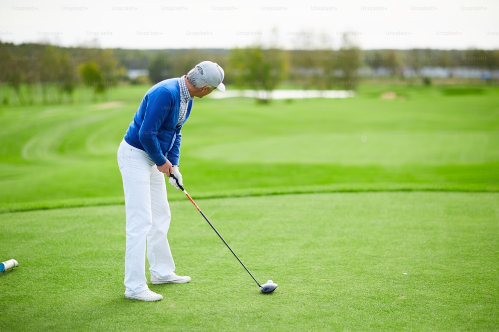 Aged man in blue cardigan, white pants and baseball cap preparing to hit golf ball with club