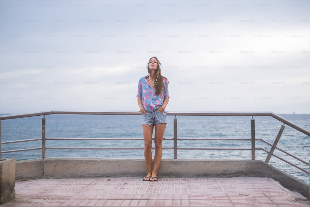 Ritratto a figura intera di bella giovane donna in piedi con l'oceano e l'acqua blu sullo sfondo e il cielo che ascolta musica con le cuffie. concetto moderno e attività di svago per la ragazza simpatica in spiaggia