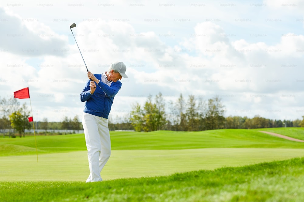 Aktiver reifer Mann, der Ball für Schläger schlägt, während er in der Freizeit auf großer grüner Wiese Golf spielt