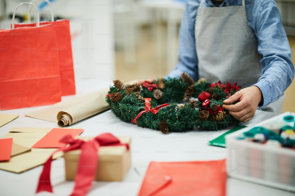 앞치마를 입은 알아볼 수 없는 남자가 종이를 들고 테이블에 서서 작업장에서 크리스마스 장식을 준비하는 동안 휴일 화환을 만드는 클로즈업