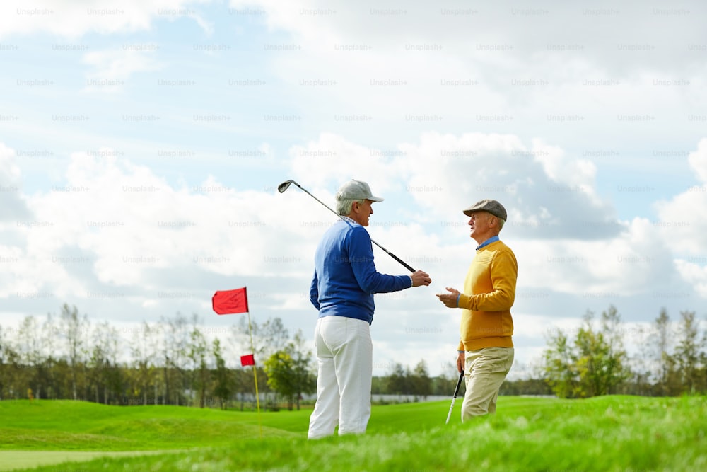 Dos hombres activos y amigables con palos de golf discutiendo el próximo juego en una nueva área de juego