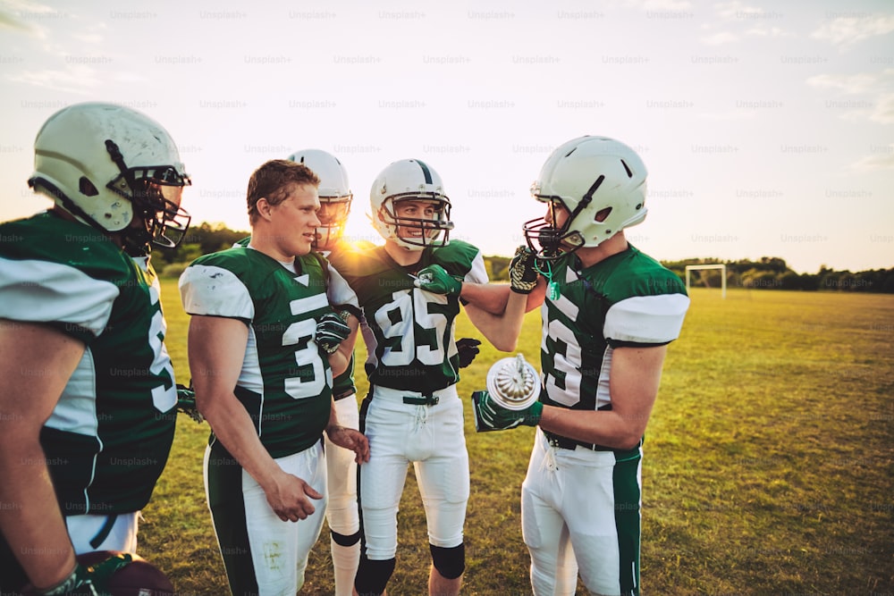 Gruppe lächelnder junger American-Football-Spieler, die zusammen auf einem Feld stehen und eine Meisterschaftstrophäe halten