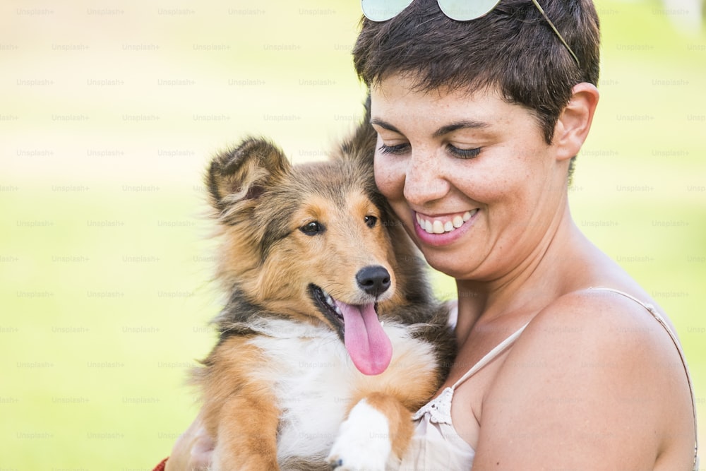 Cheerful young beautiful short black hair lady hug and take a nice Shetland puppy pet dog. Romance and friendship concept with green natural defocused bokeh background