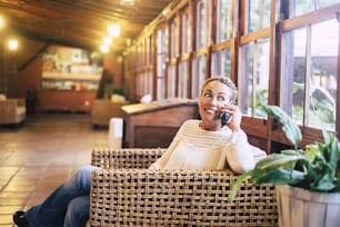 woman sitting on wicker chair in wooden house with large picture window smiling speaking on cellphone and looking outside. Home leisure activity concept for attractive middle age lady