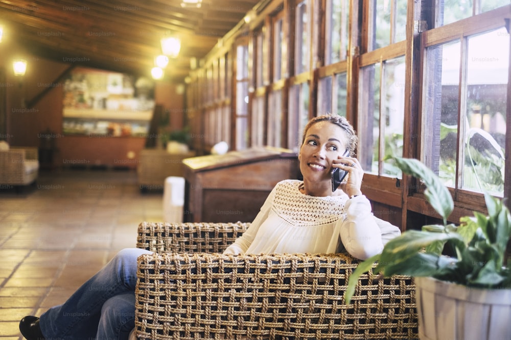 woman sitting on wicker chair in wooden house with large picture window smiling speaking on cellphone and looking outside. Home leisure activity concept for attractive middle age lady