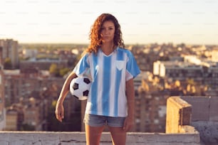 Young woman wearing a football jersey standing on a building rooftop, holding a ball and watching a sunset over the city.