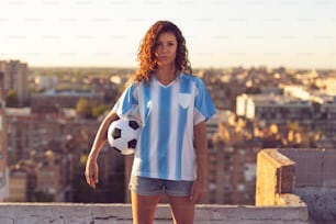 Mujer joven con una camiseta de fútbol de pie en la azotea de un edificio, sosteniendo una pelota y mirando una puesta de sol sobre la ciudad.