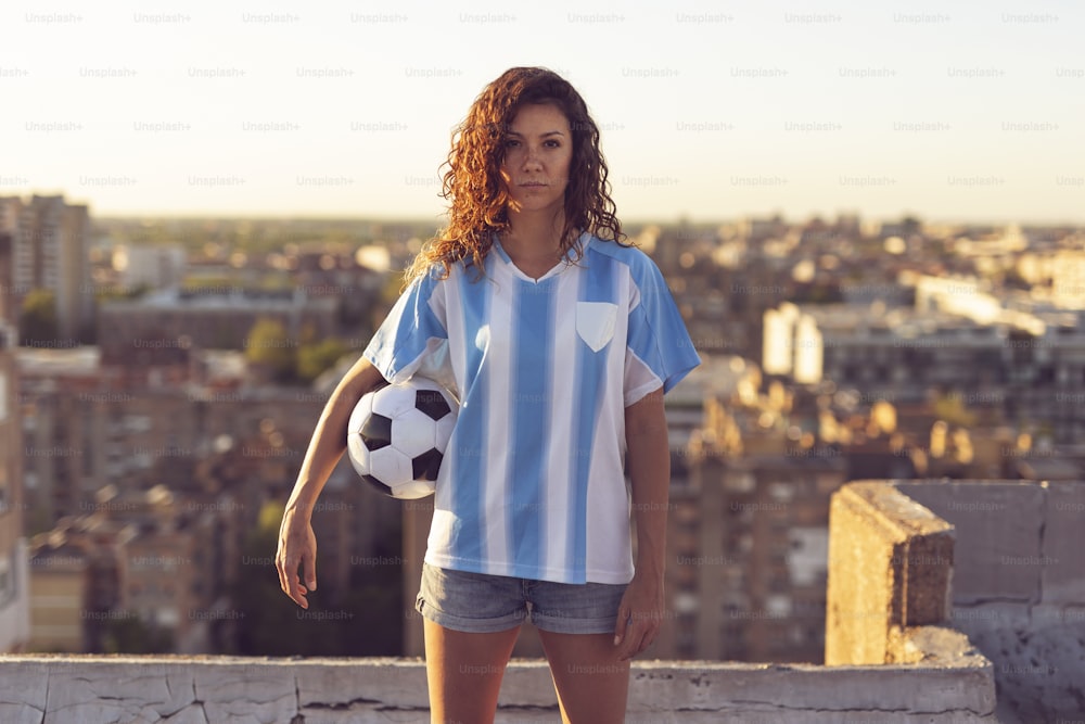 サッカーのジャージを着た若い女性がビルの屋上に立ち、ボールを持ち、街に沈む夕日を眺めている。