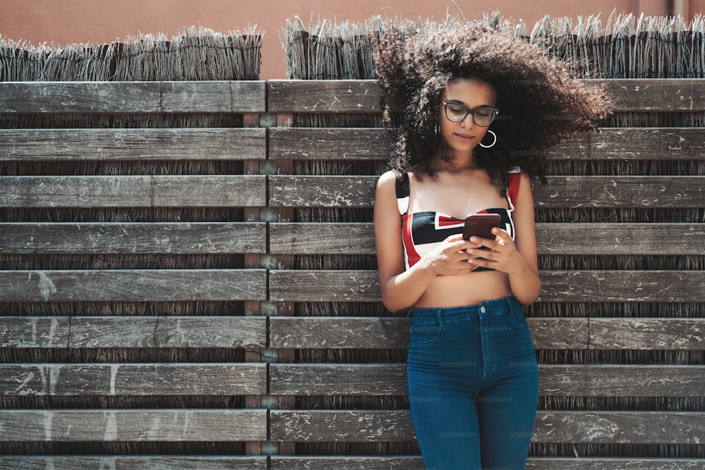 Charming young biracial female leaning against a wooden hedge with wattle fence inside and checking incoming messages on her smartphone, with a copy space area on the left for your text or logo