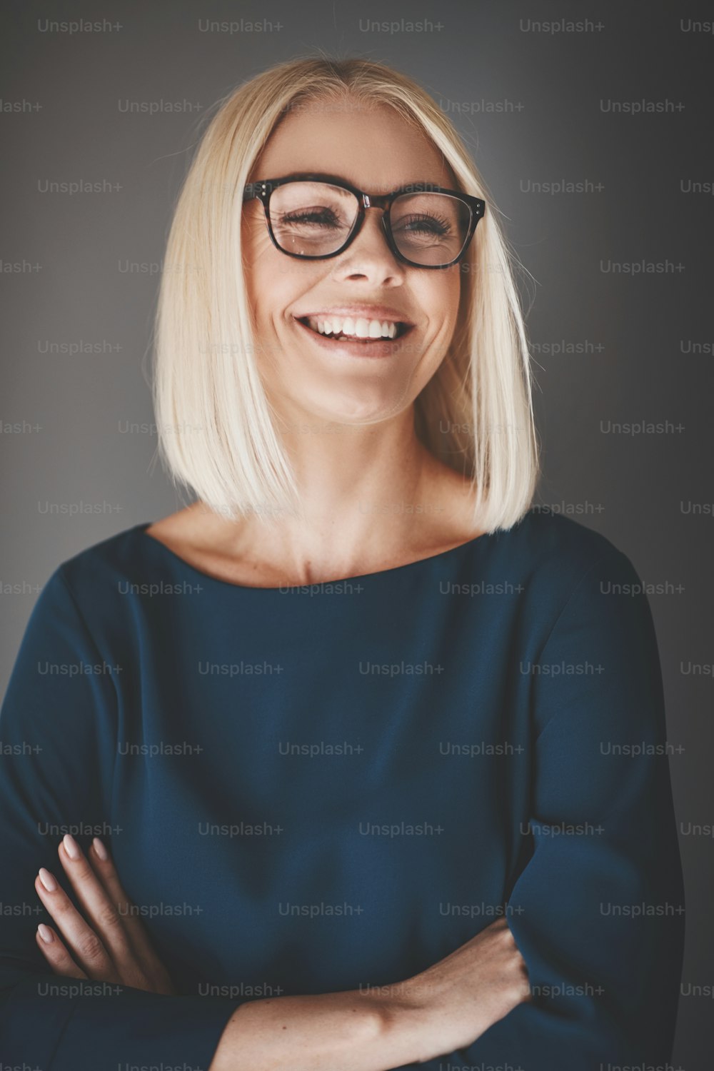 Jovem empresária sorridente usando óculos de pé com os braços cruzados contra um fundo cinza
