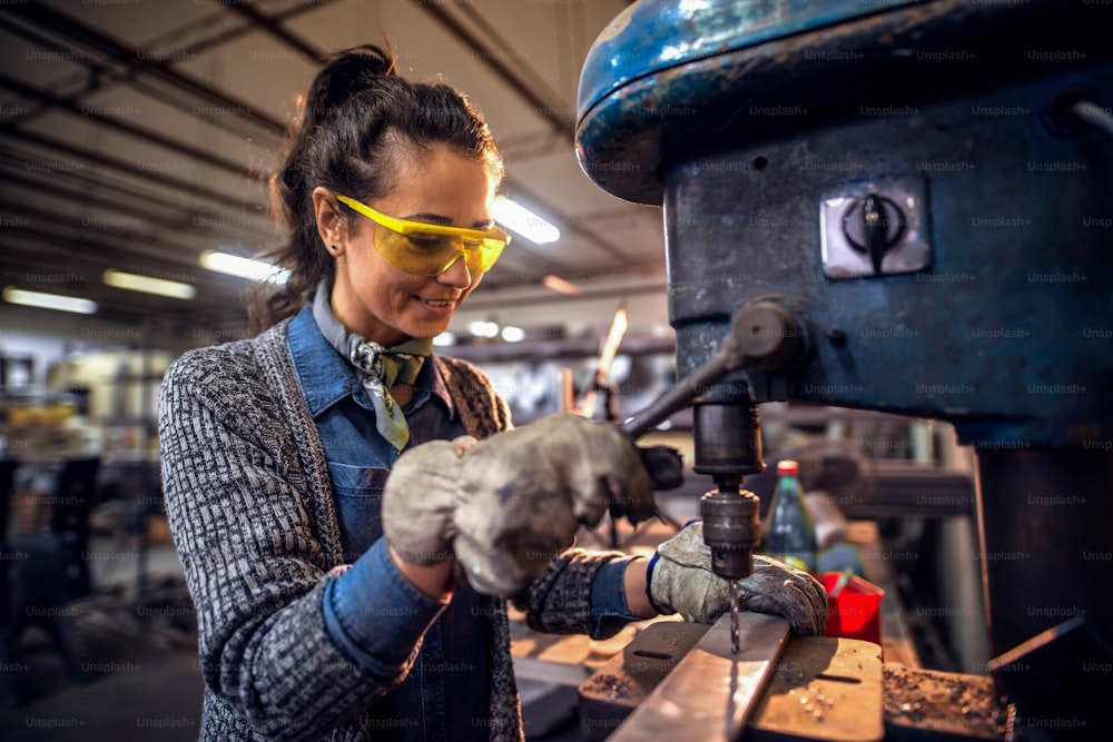 Woman using a vertical boring  machine to make a hole in metal while standing in workshop.